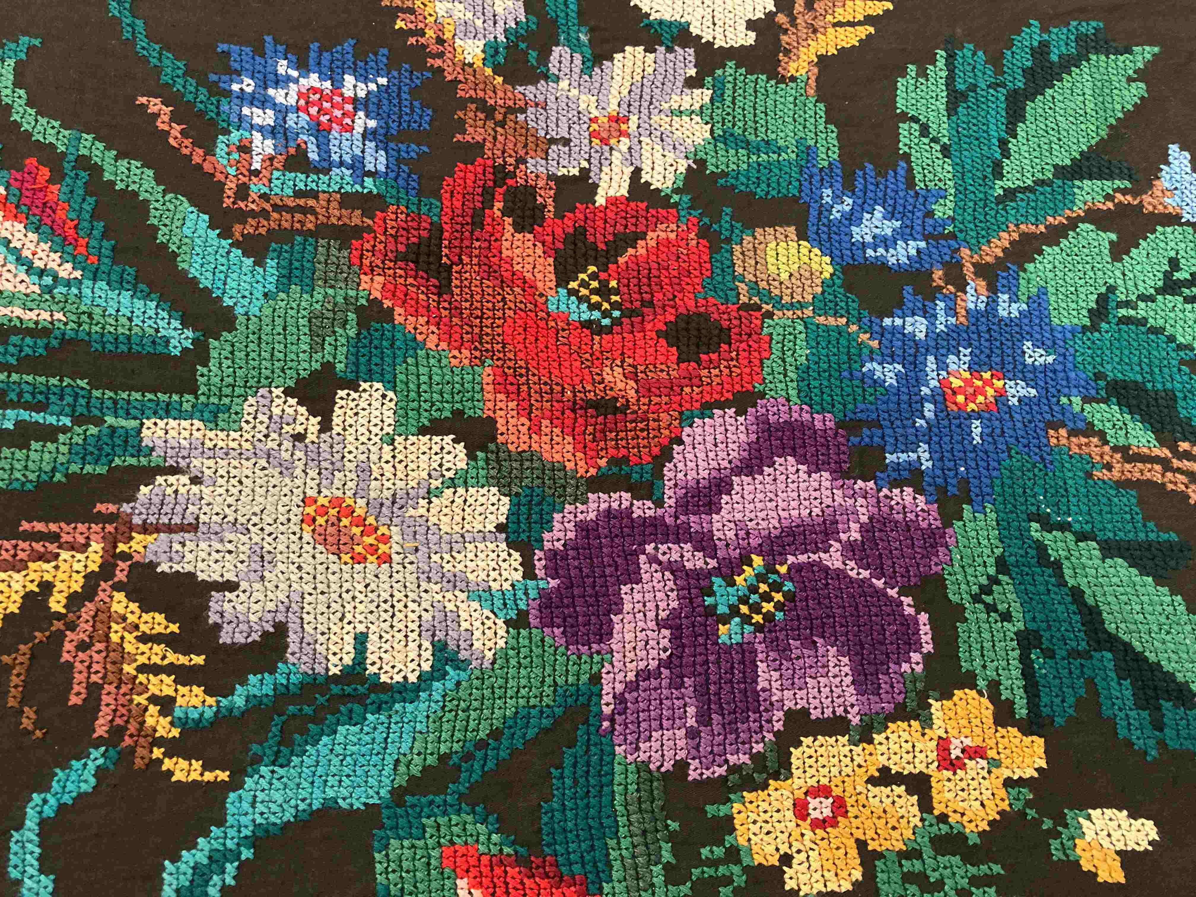 Modern cross stitch embroidery — vibrant floral bouquet with red, purple, blue, and yellow flowers on dark fabric, X-shaped stitches in embroidery hoop